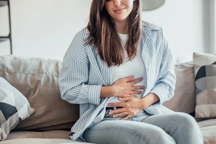 Młoda kobieta siedzi na kanapie, trzyma dłonie na brzuchu
