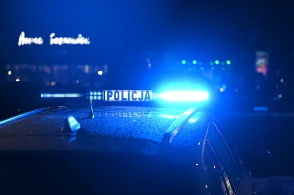 Policyjny radiowóz z włączonym niebieskim światłem stoi nocą na ulicy przed oświetlonym budynkiem.