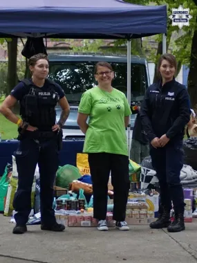 Dwie policjantki i kobieta w zielonej koszulce stoją przed namiotem z artykułami spożywczymi i zoologicznymi podczas zbiórki.