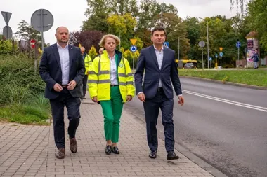 Trzy osoby, w tym dwie w garniturach i jedna w żółtej kurtce odblaskowej, idą chodnikiem wzdłuż ulicy w otoczeniu zieleni i znaków drogowych.